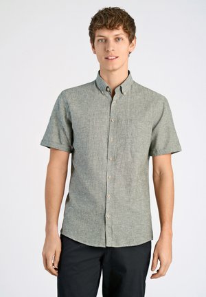 Camisa de manga corta con botones en una tela texturizada verde claro, con un bolsillo en el pecho y botones beige. Combinada con pantalones oscuros.