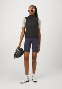 Gilet de cyclisme sans manches noir, t-shirt à manches courtes gris clair, shorts navy, chaussures de cyclisme blanches, casque noir et lunettes de soleil miroir.