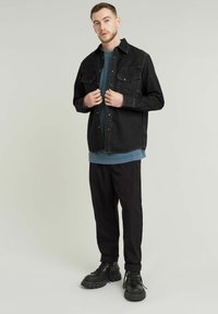 Camisa de mezclilla negra con dos bolsillos en el pecho, llevada sobre una camisa de manga larga gris-azulada, combinada con pantalones oscuros ajustados y botas negras con cordones.