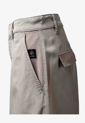 Detailní záběr béžových kalhot typu cargo s kapsou s průstřihem, klopovou kapsou, poutky na opasek a černou značkovou etiketu Elias Rumelis.