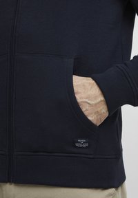 Veste à capuche zippée marine en tissu doux, dotée d'une poche kangourou, d'un ourlet côtelé et d'une étiquette cousue en bas à gauche.