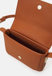 Braune Tasche aus strukturiertem Leder mit offenem Überschlag und magnetischem Druckverschluss. Innen mit Kartenfach und glattem Futter. Verstellbarer Riemen.