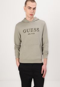 Hellgrüner Hoodie mit "GUESS EST. 1981" in einer dunkleren Farbe bedruckt. Verfügt über eine Kängurutasche und gerippte Bündchen. Kombiniert mit schwarzen Hosen.