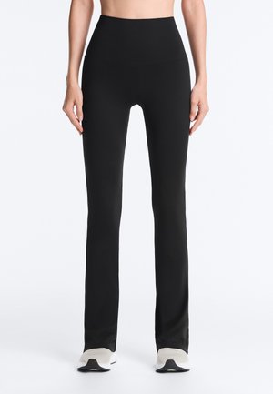 Femeie purtând pantaloni negri mulați, cu talie înaltă și ușor evazați la bază, și adidași albi, stând în picioare pe un fundal simplu.