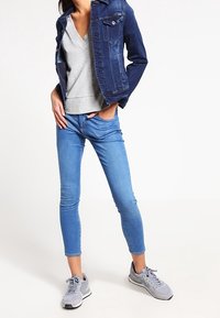 Dunkelblaue Jeansjacke mit silbernen Beschlägen, grauer V-Ausschnitt-Pullover, hellblaue Jeans und graue Sportschuhe. Schlanke Passform und lässiges Design.
