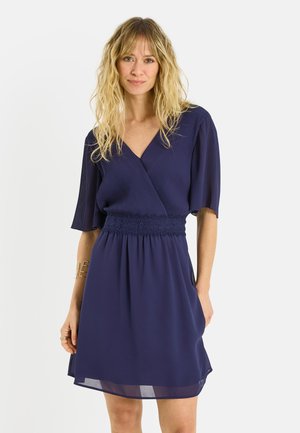 Femme aux cheveux blonds ondulés portant une robe bleu marine longueur genou avec des manches courtes plissées et une ceinture en dentelle, debout devant un fond uni.