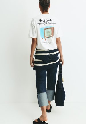 Personne portant un T-shirt blanc avec un graphique de télévision vintage et du texte, un pull rayé noué autour de la taille, un jean retroussé, tenant un sac foncé.