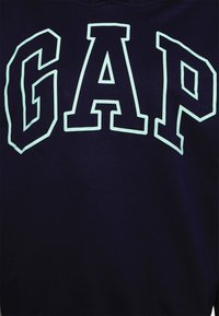 Mörkblå sweatshirt med stort vitt "GAP"-textlogo med en svag glödande effekt. Materialet verkar mjukt med en slät textur.