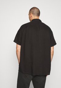 Maglietta nera a maniche corte realizzata in lino, con vestibilità comoda, colletto a punta e orlo dritto. La vista posteriore mostra una texture liscia.