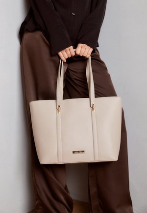 Person iført brune bukser og sort skjorte holder en beige lædertaske med to håndtag og gyldne detaljer.