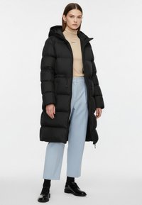 Lang sort vatteret frakke med hætte, med et vandret quiltet design. Kombineret med lyseblå bukser og sorte sko under en beige top.