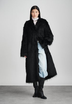 Black faux fur coat with long sleeves, open front, and textured finish, paired with light blue jeans and black knee-high boots.