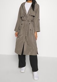 Trench coat lungo di colore marrone chiaro con cappuccio, bottoni doppi e cintura in vita. Indossato su una camicia bianca e pantaloni neri, abbinato a sneakers bianche.