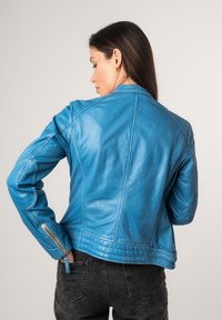Blaue Lederjacke mit taillierter Passform, hochkragen, sichtbaren Nähten und Reißverschluss-Manschetten. Die strukturierte Oberfläche verstärkt die optische Anziehungskraft.