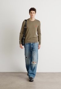 Olivenfarget genser, løse blå jeans med slitte hull, og svarte sko, accessorert med en svart skulderveske.