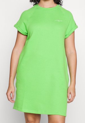 Osoba nosząca jasnozieloną, casualową sukienkę z krótkim rękawem, podwiniętymi mankietami i małym białym napisem na piersi i rękawie.