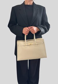 Beige kožená kabelka se strukturovaným designem, dvěma horními ušima a zlatou sponou; držena osobou v černém oversized blejzru.