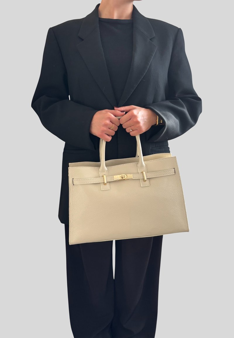 Beige kožená kabelka se strukturovaným designem, dvěma horními ušima a zlatou sponou; držena osobou v černém oversized blejzru.