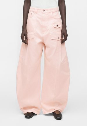 Wide-leg light pink pants with multiple buttoned flap pockets worn with black square-toe shoes and a white top.