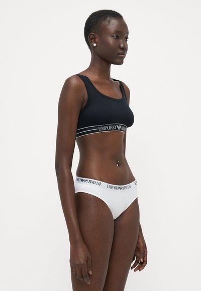 Fekete sportmelltartó fehér Emporio Armani logós pánttal; párosítva fehér bikinialsóval, amelynek logós derékpántja van. Sima szövet, elegáns dizájn.