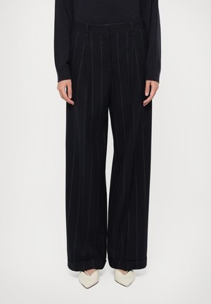 Svarte vide bukser med vertikale hvite stripes, laget av glatt stoff, har en skreddersydd midje og en oppbrett på kanten.