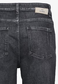 Jeans de mezclilla negra con una cintura de media altura, bolsillos tipo parche y una etiqueta de marca en color marrón cosida en la parte posterior de la cintura.