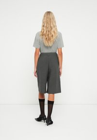 Chemise grise à manches courtes associée à des culottes grises à taille haute et à motifs. Le modèle porte des chaussettes noires et des talons, vu de dos.
