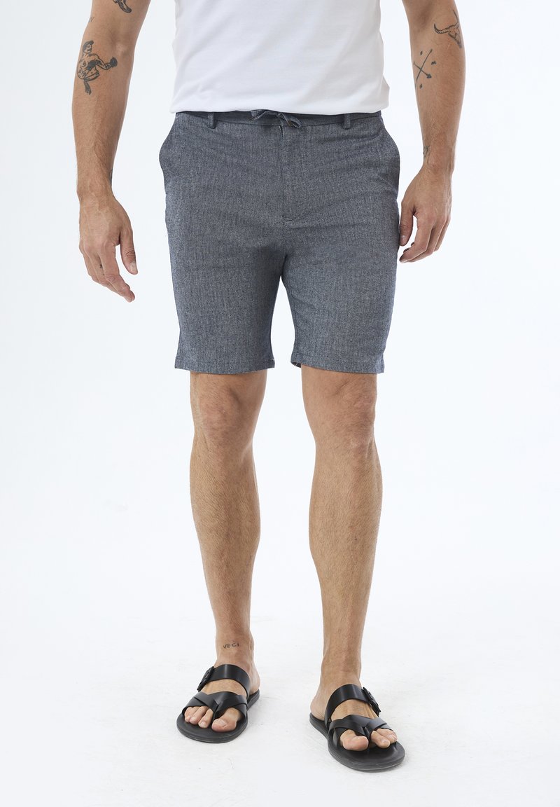 Shorts gris texturés pour hommes avec une taille à cordon, arrivant juste au-dessus du genou ; portés avec des sandales plates noires ; sans motifs ni accents visibles.
