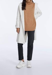 Trench-coat beige clair porté sur un pull à col roulé camel, associé à un jean foncé et des baskets blanches. Le manteau présente une taille ceinturée et des épaulettes.