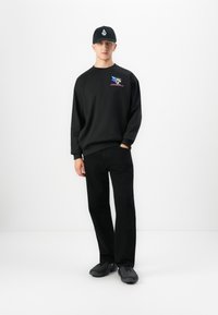 Ung man klädd i en helt svart avslappnad outfit med grafisk sweatshirt, svarta byxor, svarta sneakers och svart keps mot vit bakgrund.