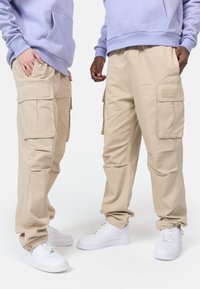 Beige cargo bukser med flere lommer, parret med hvide sneakers. Designet med en afslappet pasform; stoffet ser solidt og letvægtsud.