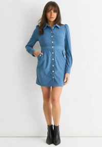 Blue denim shirt dress with long sleeves, button-down front, and side pockets. Features a fitted waist and puffed shoulder design. Black ankle boots.