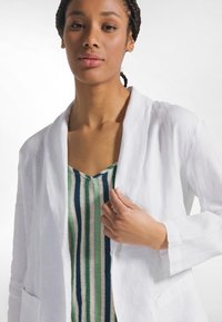 Biały lniany żakiet z kołnierzem typu szalowego i przednimi kieszeniami, noszony na zielono-niebieskim prążkowanym topie na ramiączkach z dekoltem w serek. Gładka faktura, casualowy fason.