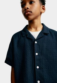 Camicia a maniche corte di un blu navy scuro con un motivo testurizzato, cinque bottoni bianchi e un colletto a rever, indossata sopra una maglietta bianca.