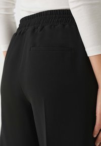 Zwarte broeken met een gladde, matte textuur, voorzien van een elastische tailleband en een enkele achterzak. Eenvoudig, functioneel ontwerp.