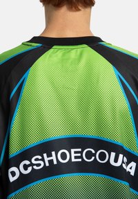 Īsā piedurkne džersija, kas izgatavota no viegla sieta auduma, ar spilgti zaļiem un melniem toņiem, ziliem akcentiem un logotipu "DCSHOECOUSA."