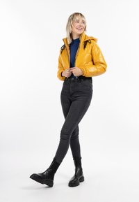 Gul glansig pufferjacka med huva och svarta dragsnören, i kombination med högmidjade svarta jeans och svarta ankelstövlar på en vit bakgrund.