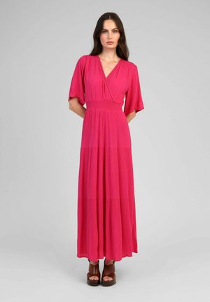 Femme debout devant un fond gris portant une robe longue rose vif à manches mi-longues et des sandales à plateforme marron.