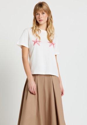 Giovane donna che indossa una t-shirt bianca a maniche corte con due stampe di stelle marine rosse e una gonna a pieghe beige a vita alta.