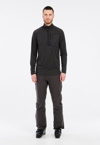 Crna dugosleeve zip-up termo majica s prednjim džepom, u kombinaciji s tamnosivim robusnim hlačama i crnim skijaškim čizmama, pogodna za zimske sportove.