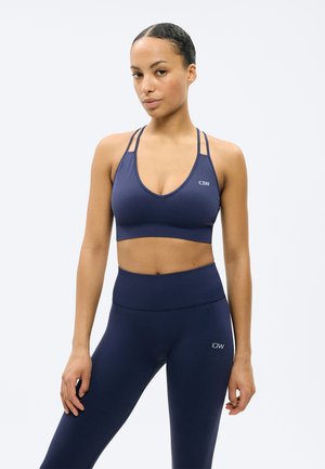 Naine, kes kannab tumesinist spordirinnahoidjat ja kõrge vöökohaga leggingse, seisab lõdvestunud poosis ühevärvilise tausta ees.