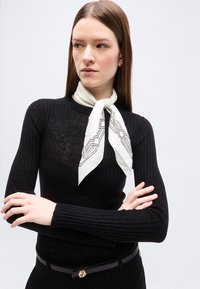 Γυναίκα με μακριά ίσια μαλλιά που φοράει μαύρο ριμπ πουλόβερ, λευκό μαντήλι με σχέδια στο λαιμό και μαύρη ζώνη, με σταυρωμένα χέρια, κοιτάζοντας πλάγια.