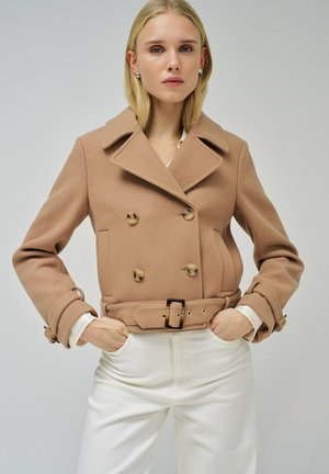 Veste marron à double boutonnage avec un large col, en tissu texturé et à taille ceinturée. Associée à un pantalon blanc, les mains dans les poches.