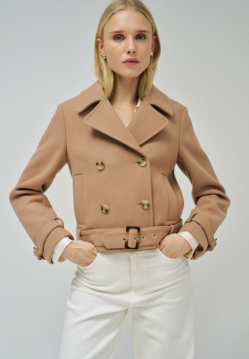 Chaqueta marrón de doble botonadura con un amplio cuello, tejido texturizado y cintura ceñida con cinturón. Combinada con pantalones blancos, manos en los bolsillos.