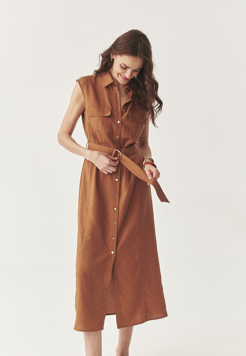 Vestido marrón sin mangas con botones en la parte delantera, dos bolsillos en el pecho, cintura con cinturón y un dobladillo de longitud media, con una textura de tela suave.