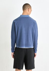Blue cotton sweatshirt with long sleeves, a short collar, and a relaxed fit, paired with black shorts. Back view shows soft texture.