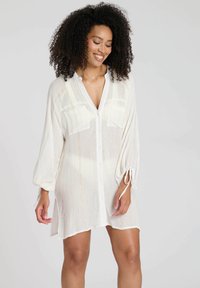 Abito camicia bianco con righe verticali, due tasche sul petto e maniche lunghe arricciate. Tessuto morbido e leggero con una leggera lucentezza.