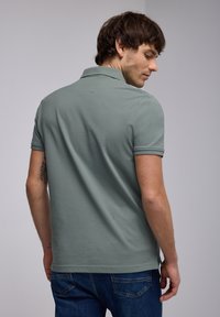 Camisa polo de algodón verde apagado con mangas cortas, presenta un cuello clásico y aberturas laterales. Lleva pantalones vaqueros de mezclilla azules.