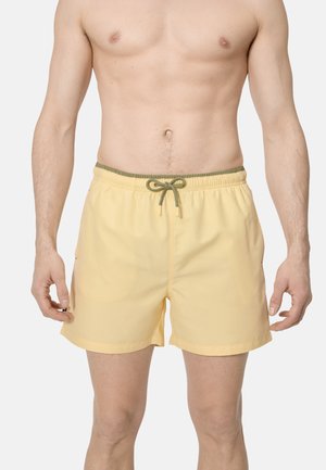 Hombre sin camiseta con pantalones cortos de baño amarillo claro con cordón y cintura de color verde oliva sobre un fondo blanco.