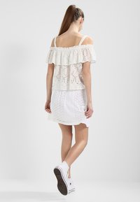Top bianco off-the-shoulder con volant sovrapposti e design in pizzo floreale. Abbinato a una gonna bianca in pizzo e scarpe da ginnastica bianche.